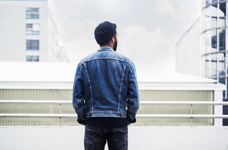 Mies seisoo selin kameraan kaupunkimaisemassa, pukeutuneena farkkutakkiin ja tummaan pipoon.