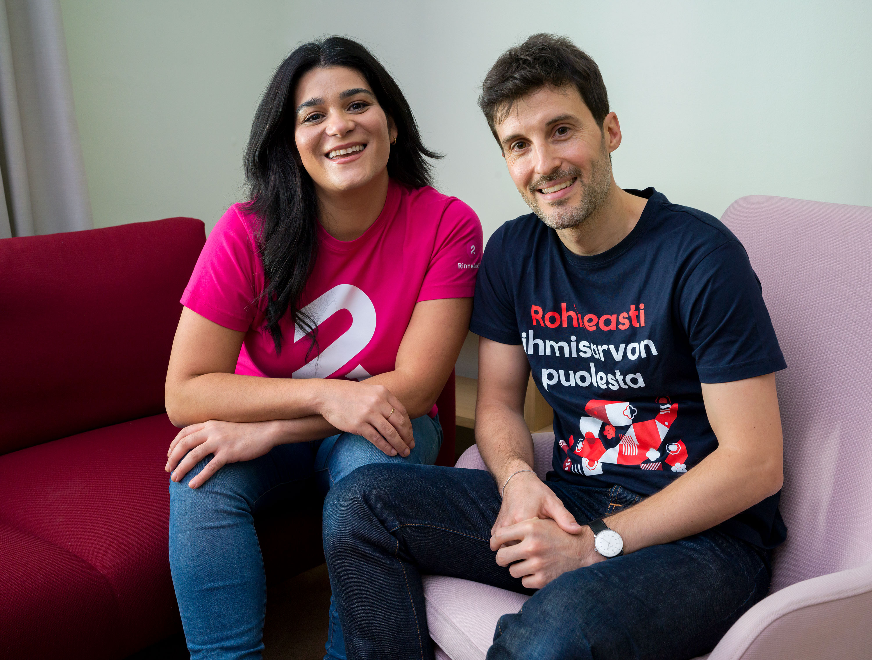 Kaksi hymyilevää henkilöä istumassa vierekkäin sohvalla, nainen pinkissä Rinnekodit-paidassa ja mies tummassa Diakonissalaitos-paidassa.