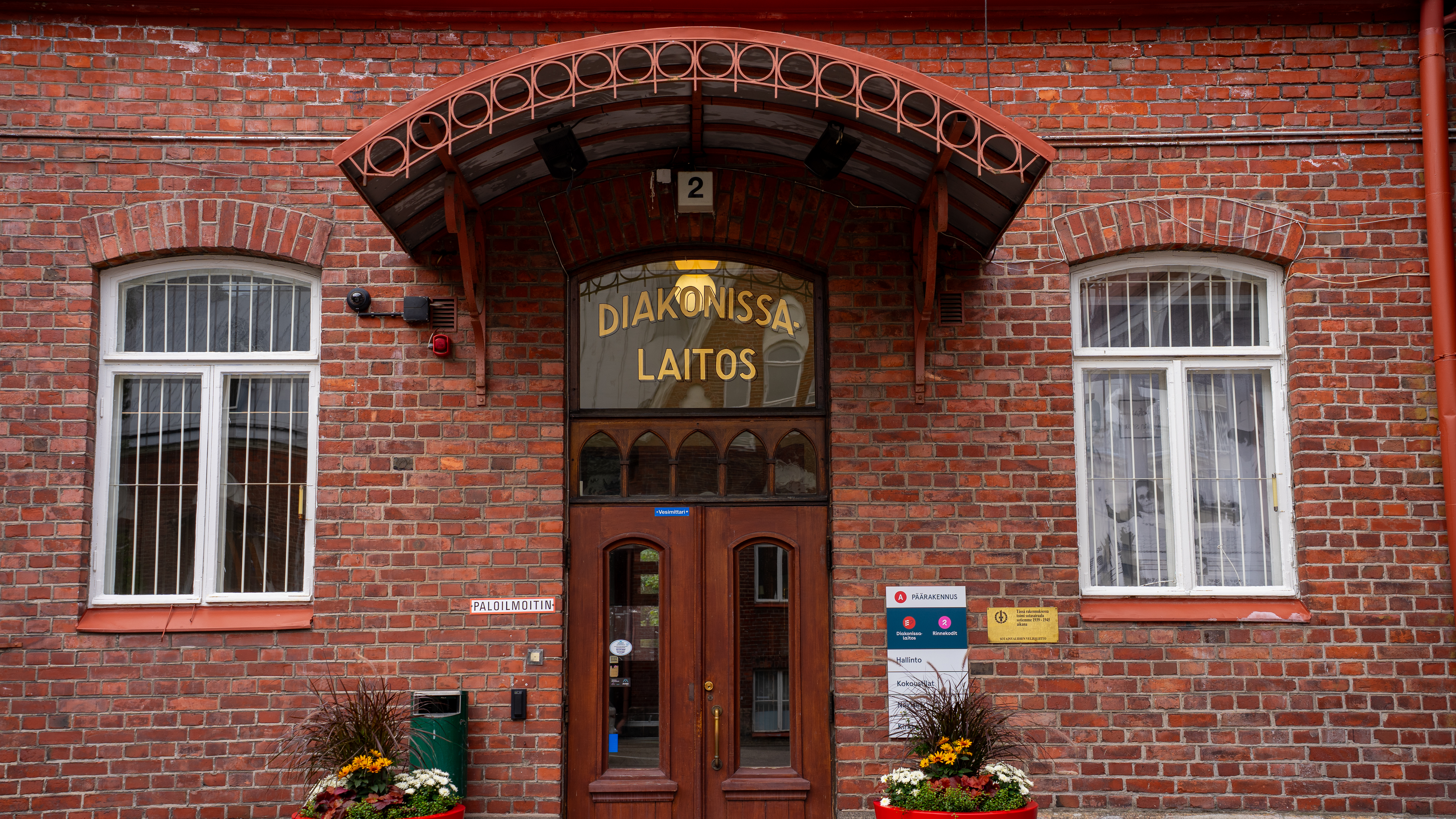 Diakonissalaitoksen punatiilinen rakennuksen sisäänkäynti, jossa ovella kyltti "Diakonissalaitos" ja kukkaistutuksia oviaukon molemmin puolin.