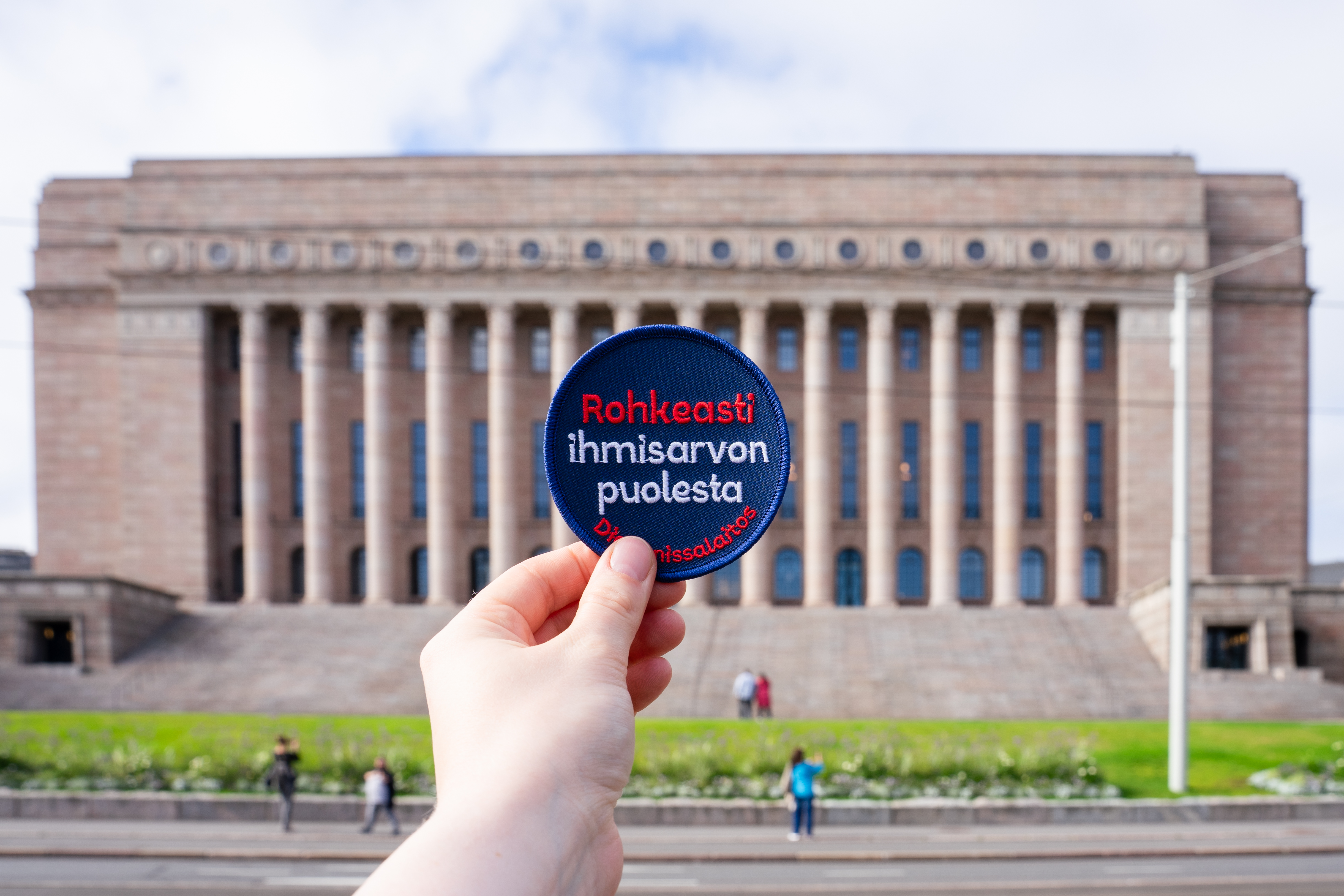 Käden pitämä pyöreä sinipunainen merkki, jossa lukee "Rohkeasti ihmisarvon puolesta", taustalla eduskuntatalo.