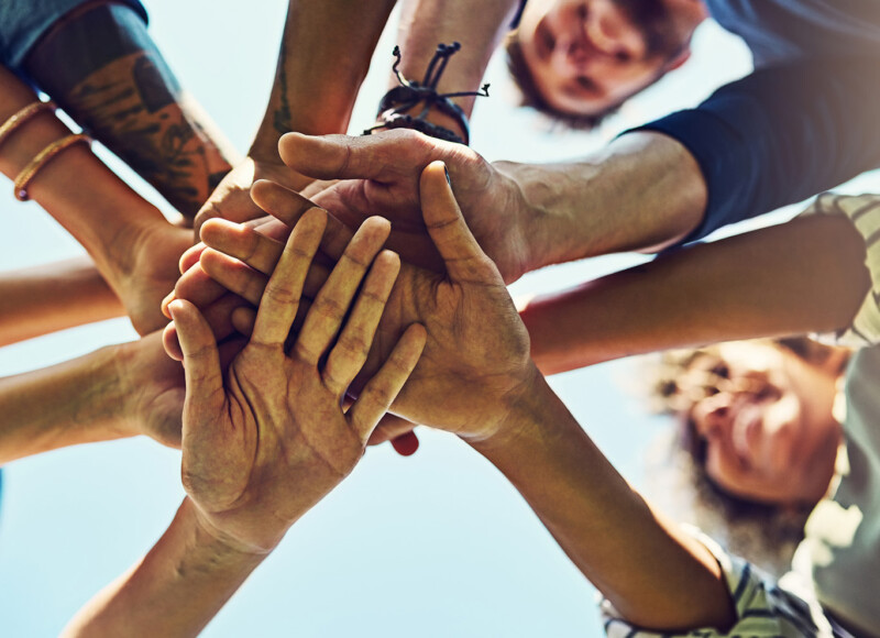 Läheltä kuvattu ryhmä ihmisiä yhdistää kätensä ulkona osoittaen yhteishenkeä ja ystävyyttä.