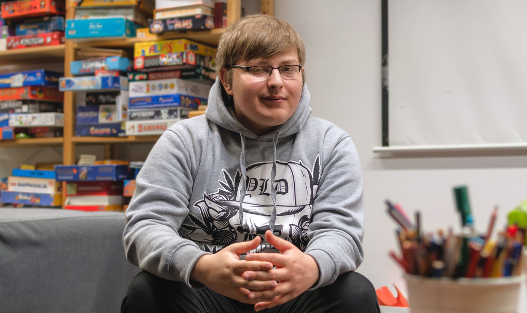 Nuori henkilö istuu kädet yhteenliitettyinä huoneessa, jossa taustalla on hylly täynnä lautapelejä.