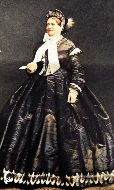 Muotokuvassa 1800-luvun puolivälin naishahmo, pukeutuneena mustaan, pitkään ja leveälahkeiseen mekkoon, jossa on valkoiset pitsikoristeet ja valkoinen huivi kaulassa, päässään koristeellinen hattu höyhenkoristeella.