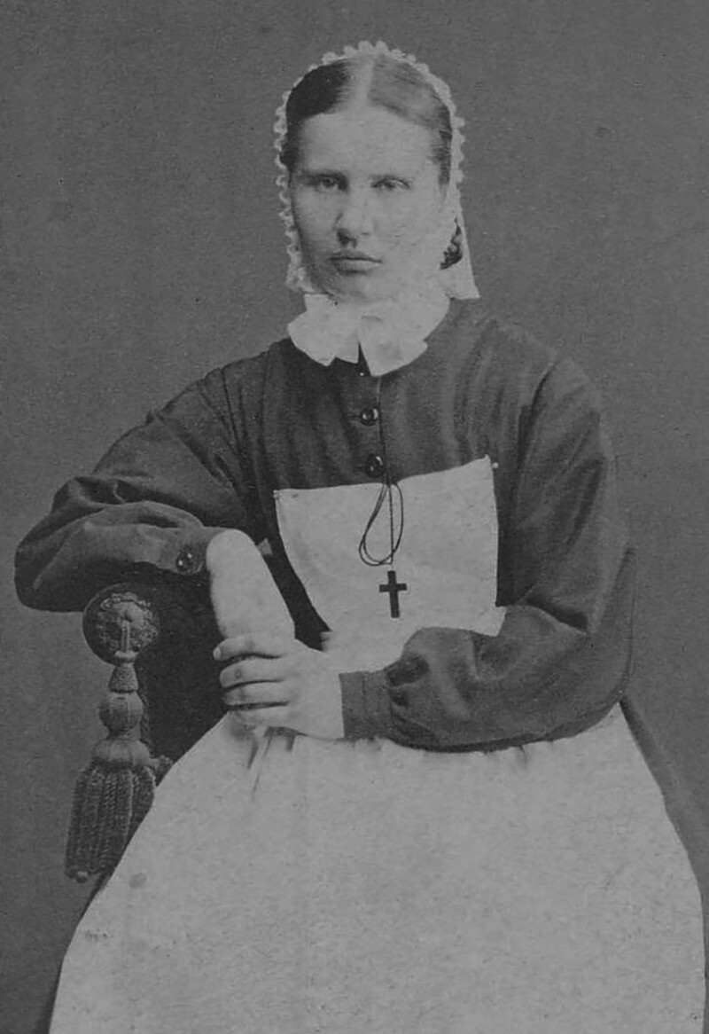 Mustavalkoinen kuva naisesta, jolla on yllään vanhanaikainen puku, valkoinen esiliina ja päässään pitsillä koristettu päähine.