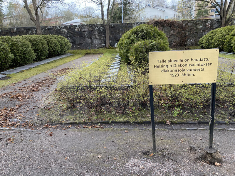 Diakonissalaitoksen diakonissojen hauta-alue, jossa kivistä muodostettu polku, pensaita ja keltainen kyltti, jossa lukee hauta-alueen perustamisesta vuodesta 1923 alkaen.