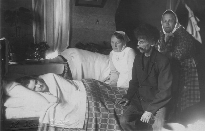 Mustavalkoinen valokuva, jossa diakonissa tekee kotikäynnin, istuu sängyn vieressä potilaan luona yhdessä kahden muun henkilön kanssa, kaikki pukeutuneina ajan mukaisiin vaatteisiin.