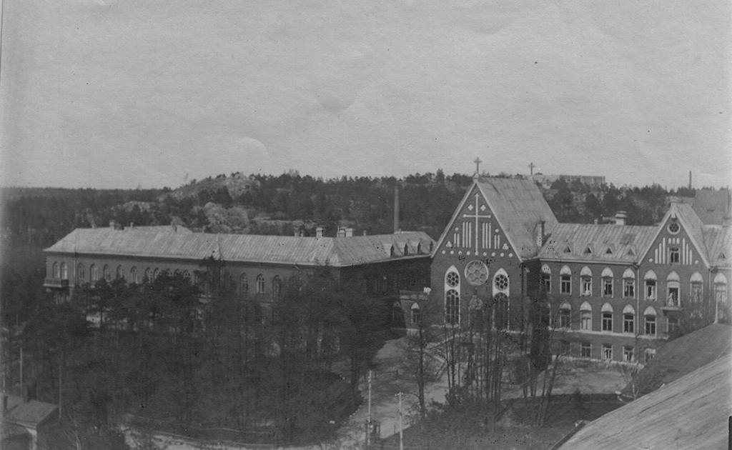 Mustavalkoinen kuva Diakonissalaitoksen kirkosta ja sen ympäristöstä, taustalla metsää ja rakennuksia.