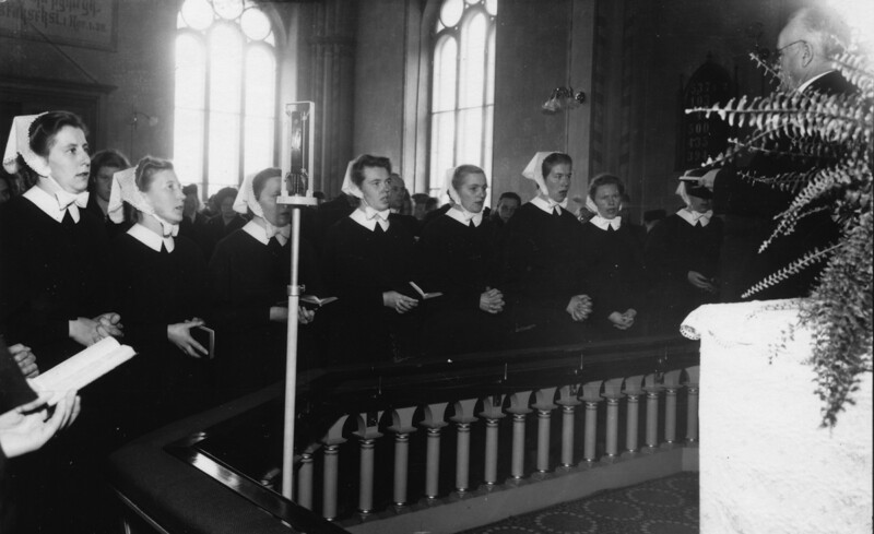 Mustavalkoinen kuva, jossa ryhmä diakonissoja istuu tai seisoo penkeissä kirkon alttarilla kuuntelemassa pappia, joka on edessä oikealla.