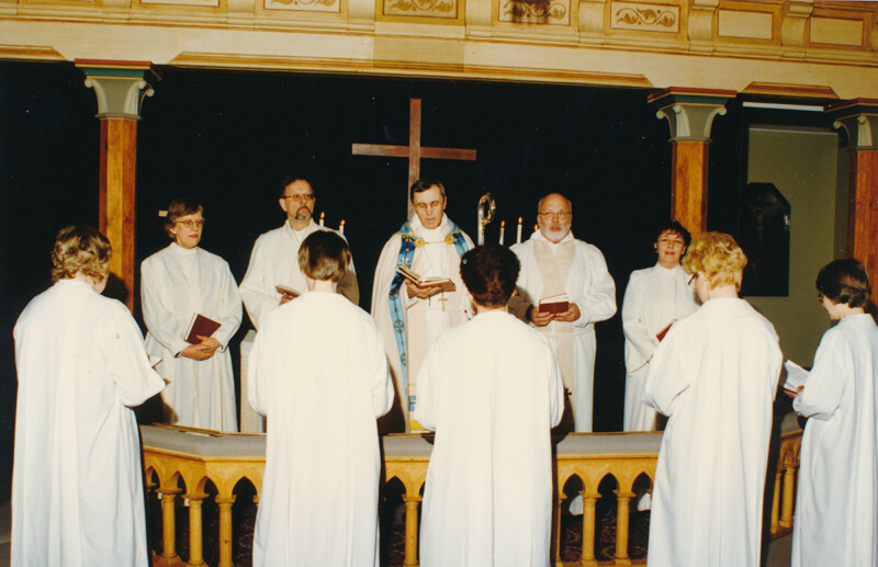 Kirkon alttarin edessä papit ja diakonissat valkoisissa liturgisissa vaatteissa jumalanpalveluksessa, taustalla suuri risti.