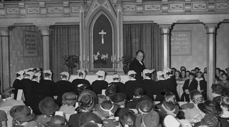 Mustavalkoinen kuva Diakonissalaitoksen kirkon alttarista vihkimistilaisuudessa vuonna 1934, jossa papisto ja naiskätilöt rivissä alttarin edessä, yleisö etualalla.
