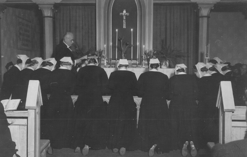 Mustavalkoinen kuva, jossa ryhmä diakonissoja polvistuneena kirkon alttarilla, pappi seisoo alttarin takana, taustalla risti ja kynttilöitä.