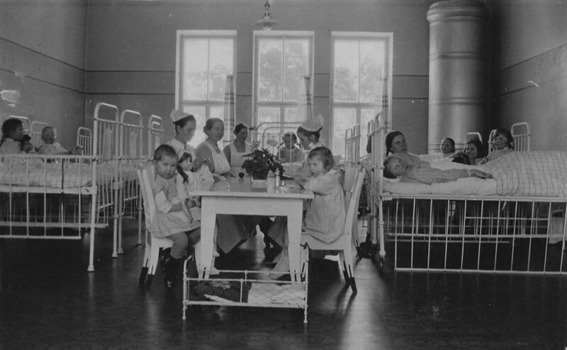 Mustavalkoinen kuva sairaalan lastenhuoneesta, jossa useita lapsia ja sairaanhoitajia istuu vuoteiden ympärillä askartelemassa ja leikkimässä.