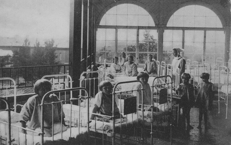 Vanha mustavalkoinen valokuva, jossa useita lapsipotilaita istuu tai makaa sairaalasängyissä avoimella parvekkeella, hoitaja seisoo heidän vieressään.