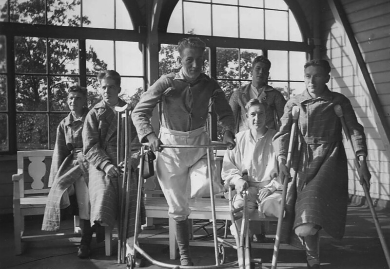 Mustavalkoinen kuva, jossa viisi nuorta miestä sairaalaympäristössä, yksi kävelee apuvälineiden kanssa, muut istuvat penkillä tai tuolilla, taustalla suuret ikkunat.