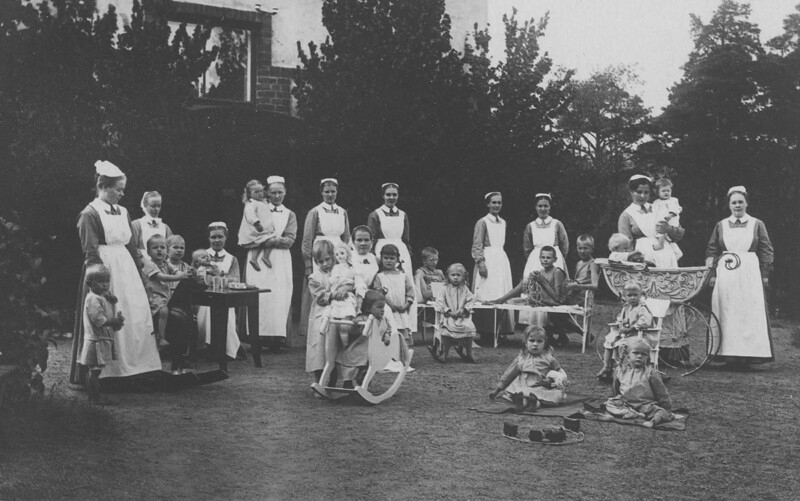 Mustavalkoinen historiallinen valokuva, jossa useita sairaanhoitajia ja lapsia ulkona puutarhassa vanhan rakennuksen edustalla.