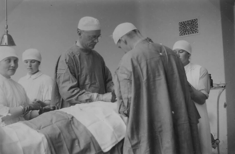 Mustavalkoinen kuva sairaalan leikkaussalista, jossa lääkärit ja hoitajat pukeutuneina kirurgisiin vaatteisiin suorittavat leikkausta potilaalle.