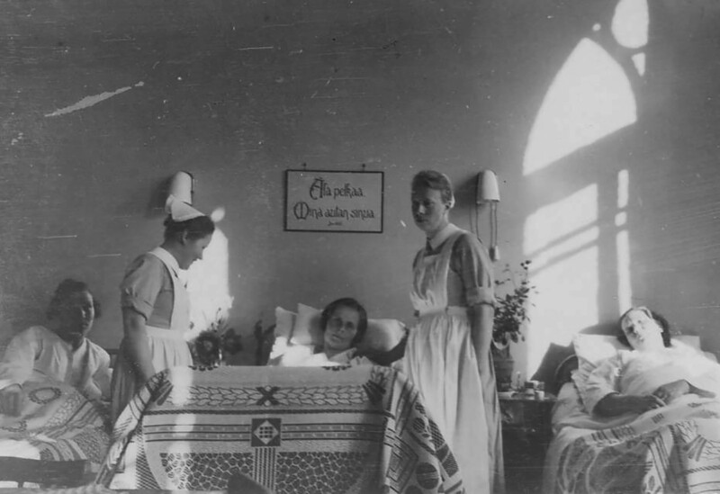 Mustavalkoinen valokuva sairaalan potilashuoneesta, jossa kaksi sairaanhoitajaa ja kolme potilasta lepäämässä vuoteissa.