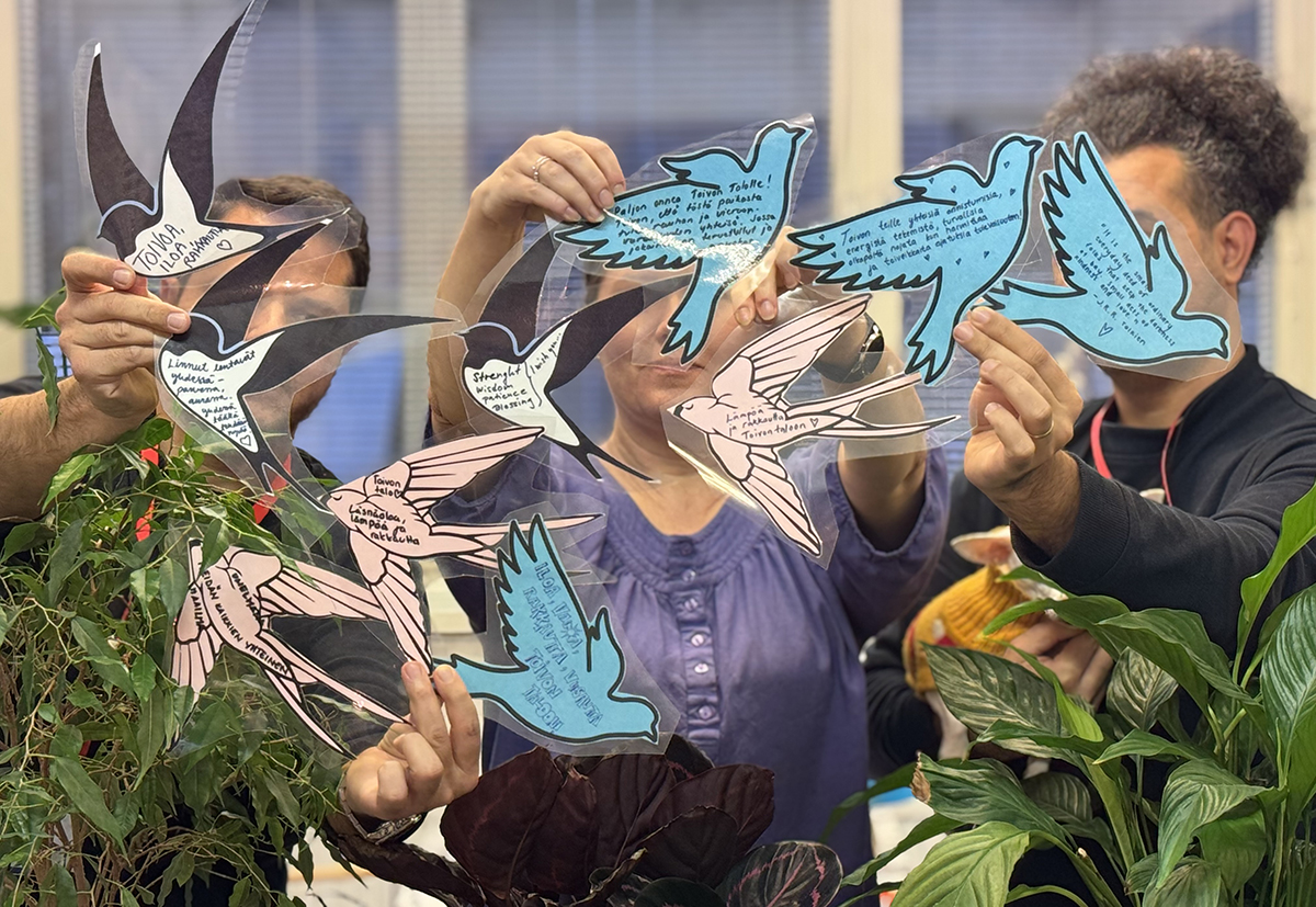 Kolme henkilöä pitelee värillisiä, paperista leikattuja lintuhahmoja, taustalla kasveja ja lasiseinä.