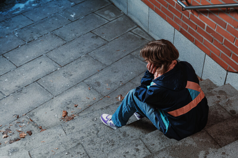 Nuori istuu yksin ulkona betonilattialla kädet poskilla, taustalla tiiliseinä.