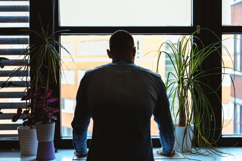 Mies nojaa ikkunalaudalle selin kameraan, ikkunan edessä huonekasveja ja taustalla rakennuksia.