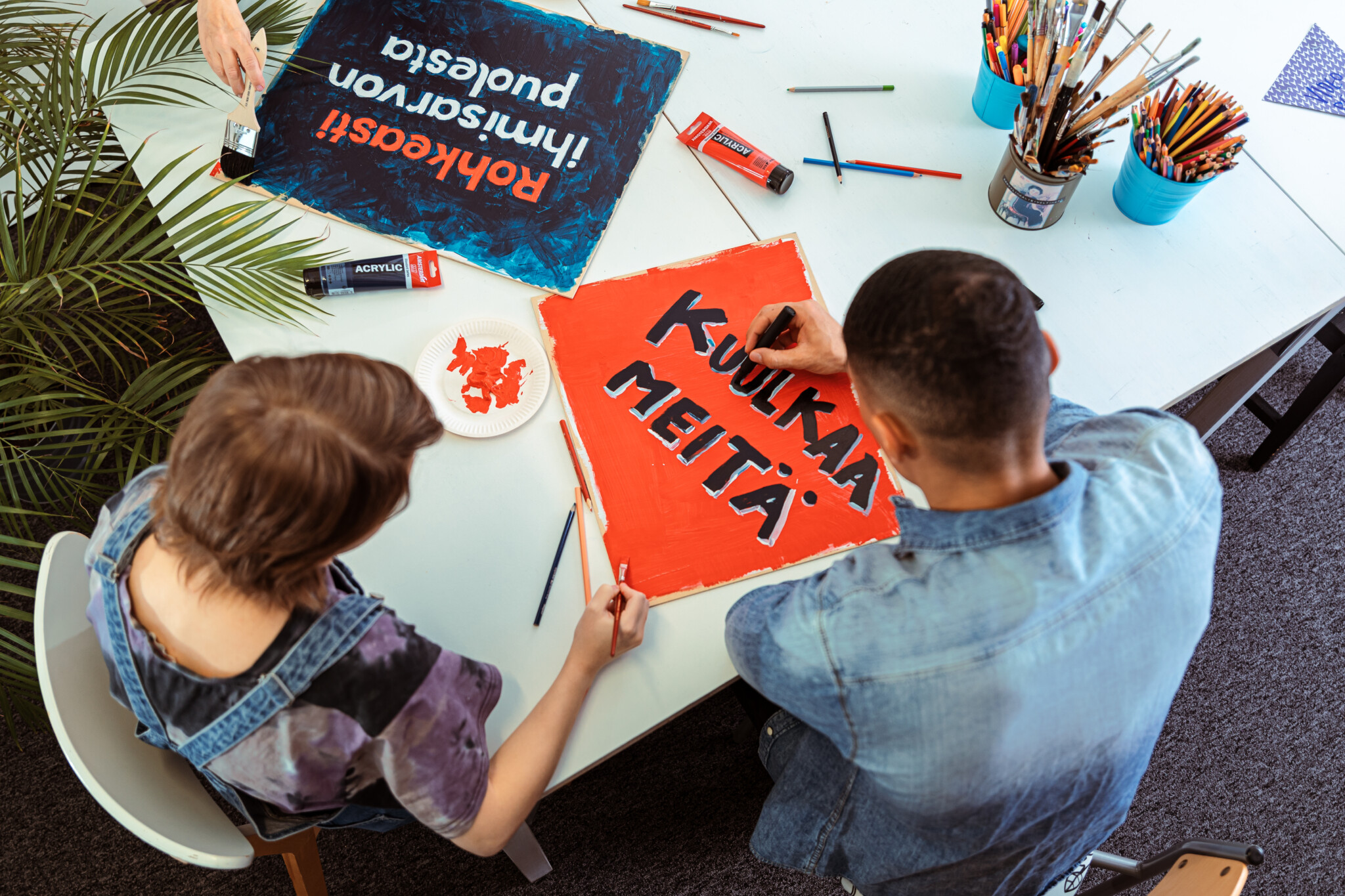 Kaksi henkilöä maalaamassa julisteita, joista yksi punainen juliste, jossa lukee "Kuulka meitä", ja toinen sininen juliste, jossa osittain näkyy teksti "Rohkeasti ihmisarvon puolesta".