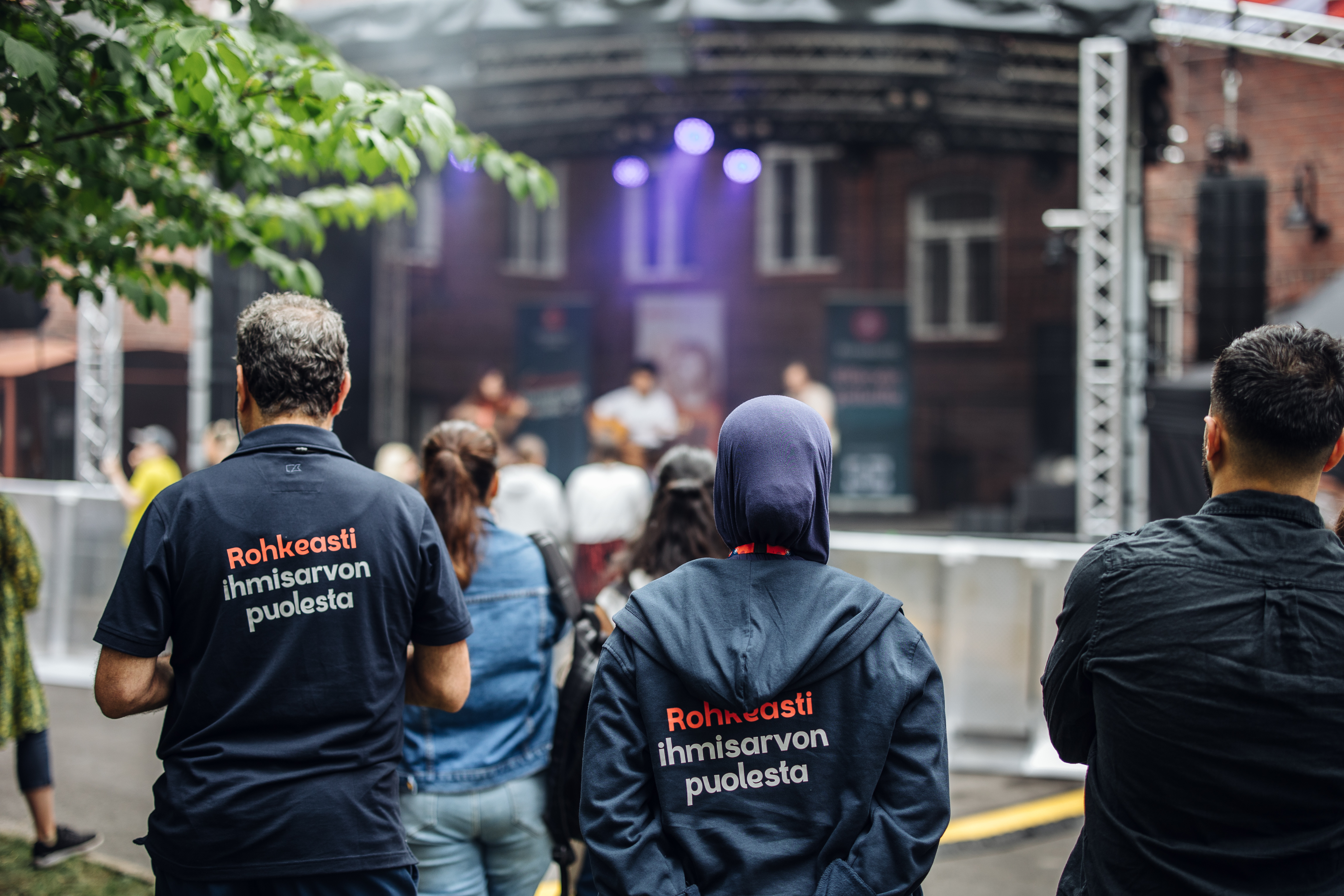 Ihmisiä katsomassa ulkoilmakonserttia, kaksi henkilöä takaa päin kuvattuna t-paidoissa joissa lukee "Rohkeasti ihmisarvon puolesta".
