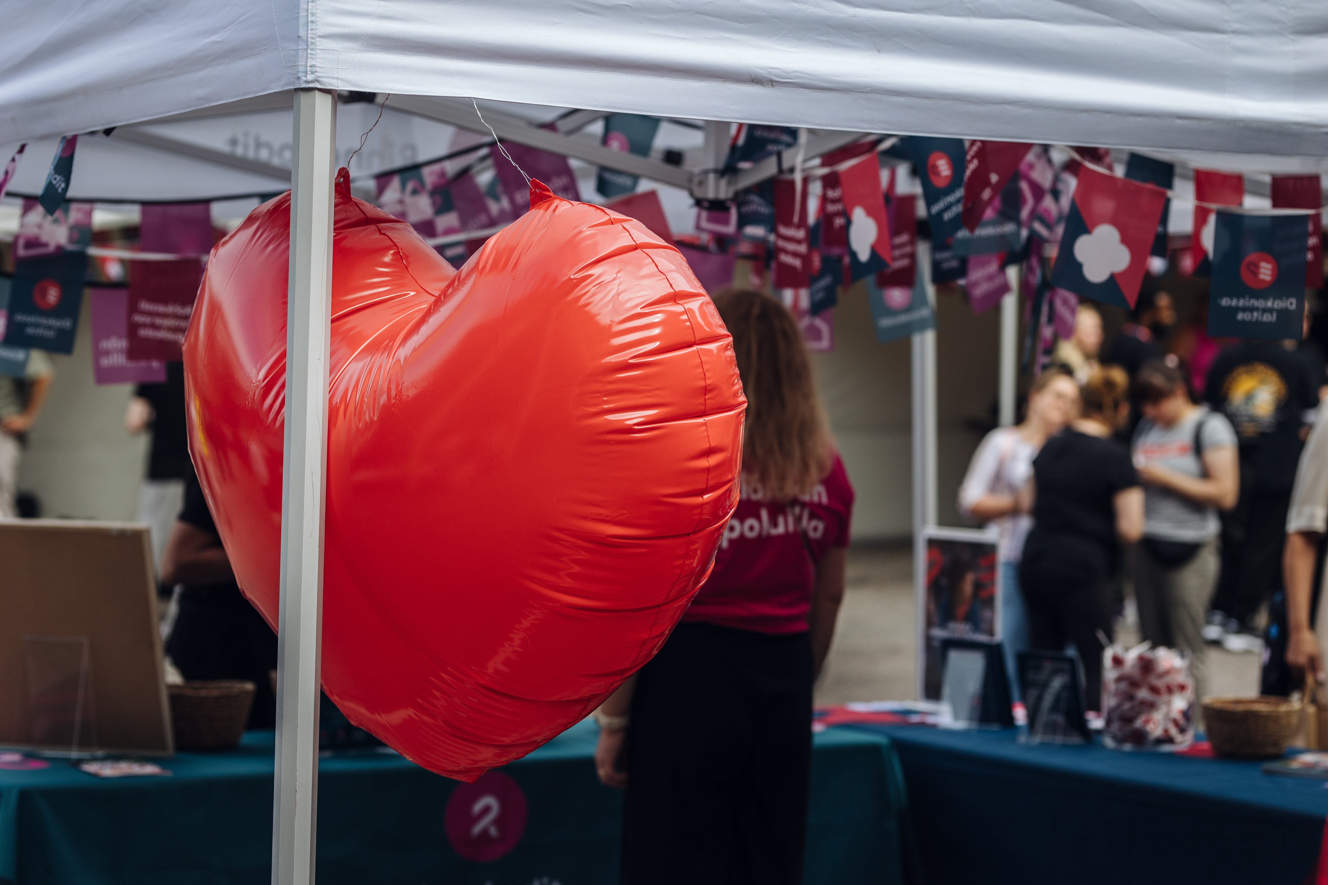 Punainen sydän-ilmapallo katoksessa, taustalla ihmisiä ja tapahtumapisteitä ulkoilmatapahtumassa.