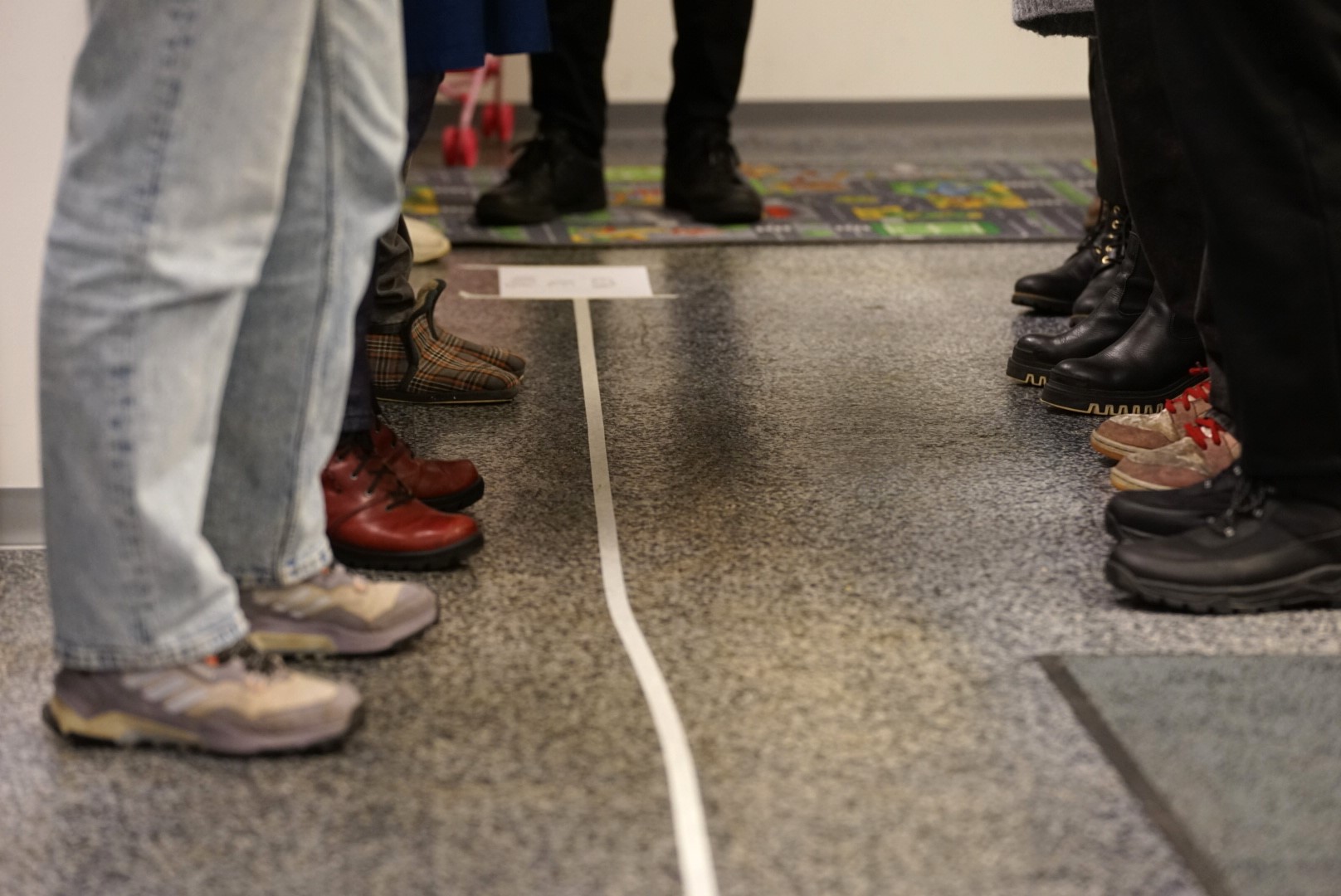 Seisovien ihmisten jalat lattialla, jossa kulkee teipattu viiva.