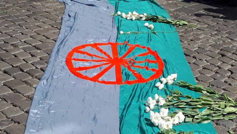 A green and blue flag with a red wheel symbol laid on cobblestone ground, adorned with white flowers placed along its edges.