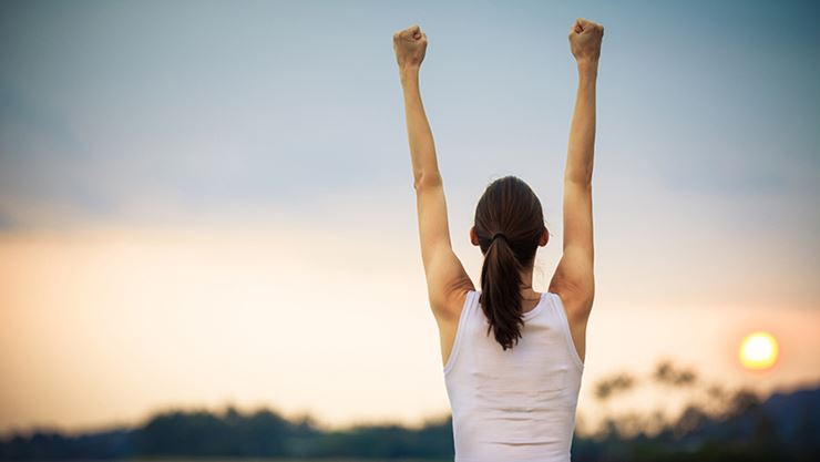 Person with ponytail raising both arms triumphantly against a sunset sky, symbolizing achievement and success.