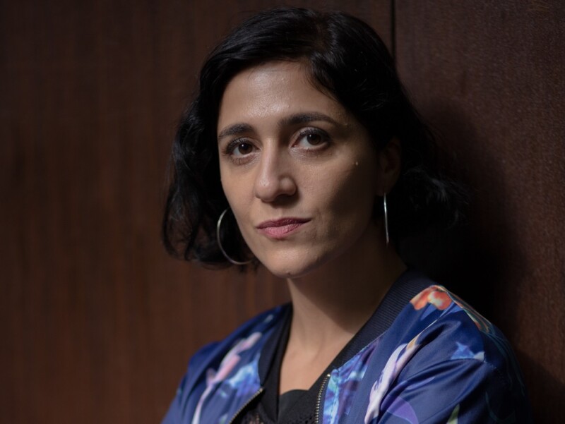 Nuori nainen mustissa hiuksissa katsoo suoraan kameraan, taustalla tumma seinä.