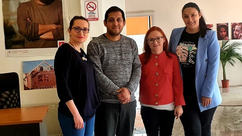 Neljä henkilöä seisoo rivissä sisätiloissa, taustalla seinällä julisteita ja kasvi.