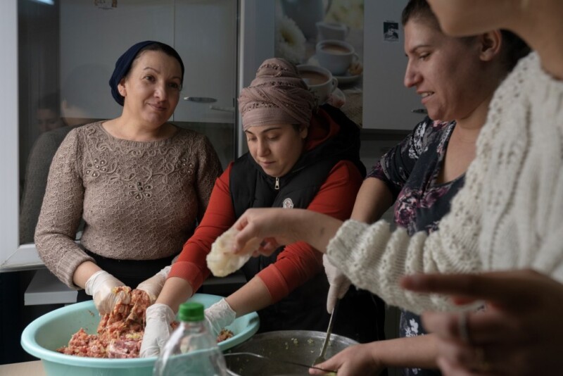 Four women gathered around a table preparing food together, mixing ingredients in bowls.
