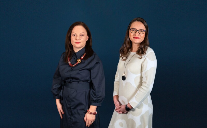 Two women standing side by side against a dark blue background, one wearing a dark blue dress and the other wearing a white dress with circular patterns, both looking at the camera.