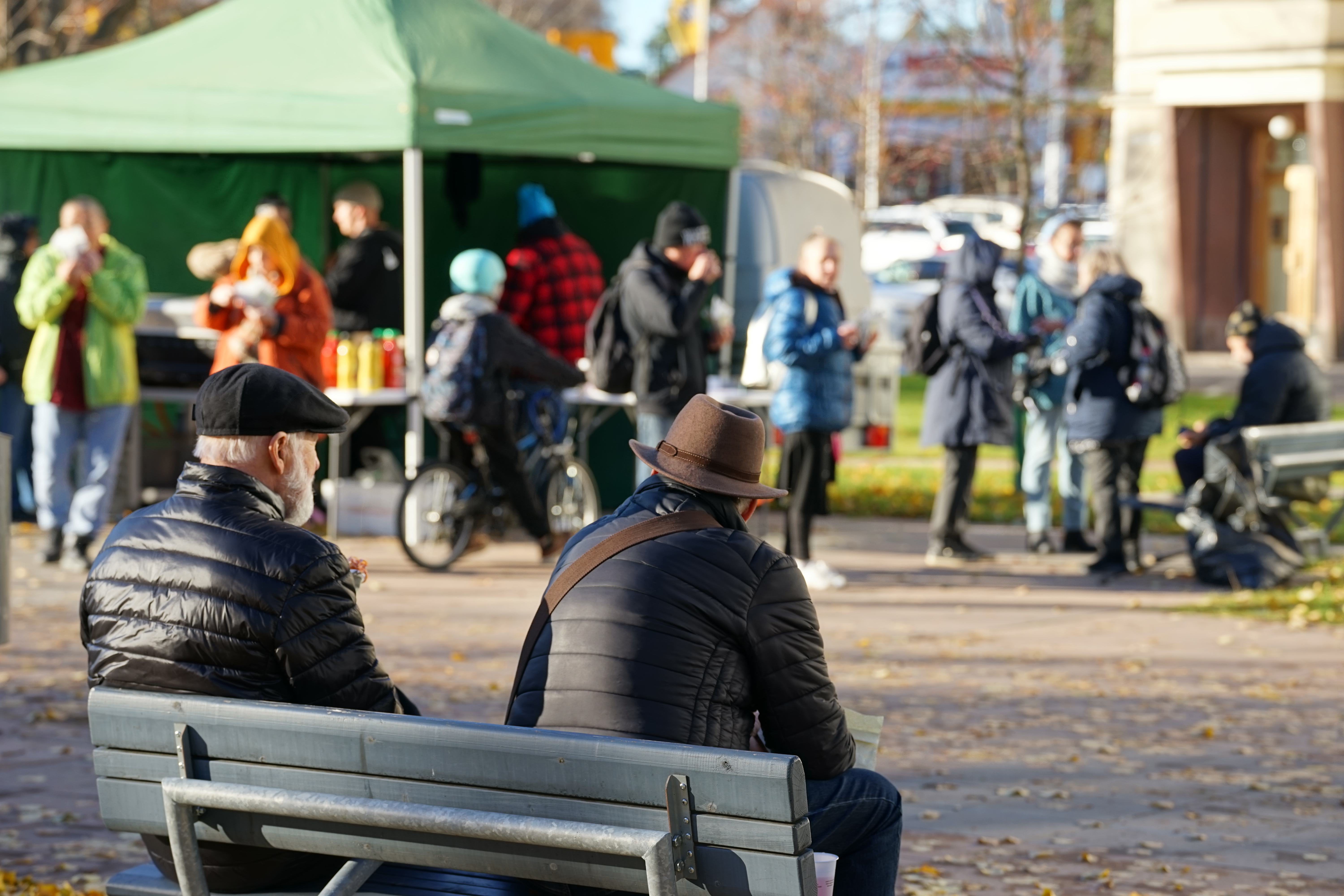 Kaksi henkilöä istuu penkillä ulkona, taustalla ihmisiä jonottamassa teltan luona syksyisessä kaupunkiympäristössä.