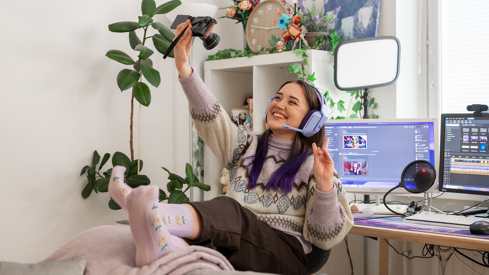 Nuori nainen istuu työpöydän ääressä pukeutuneena villapaitaan ja kuulokkeisiin, hymyilee ja pitää kameraa kädessään ottaen selfietä.