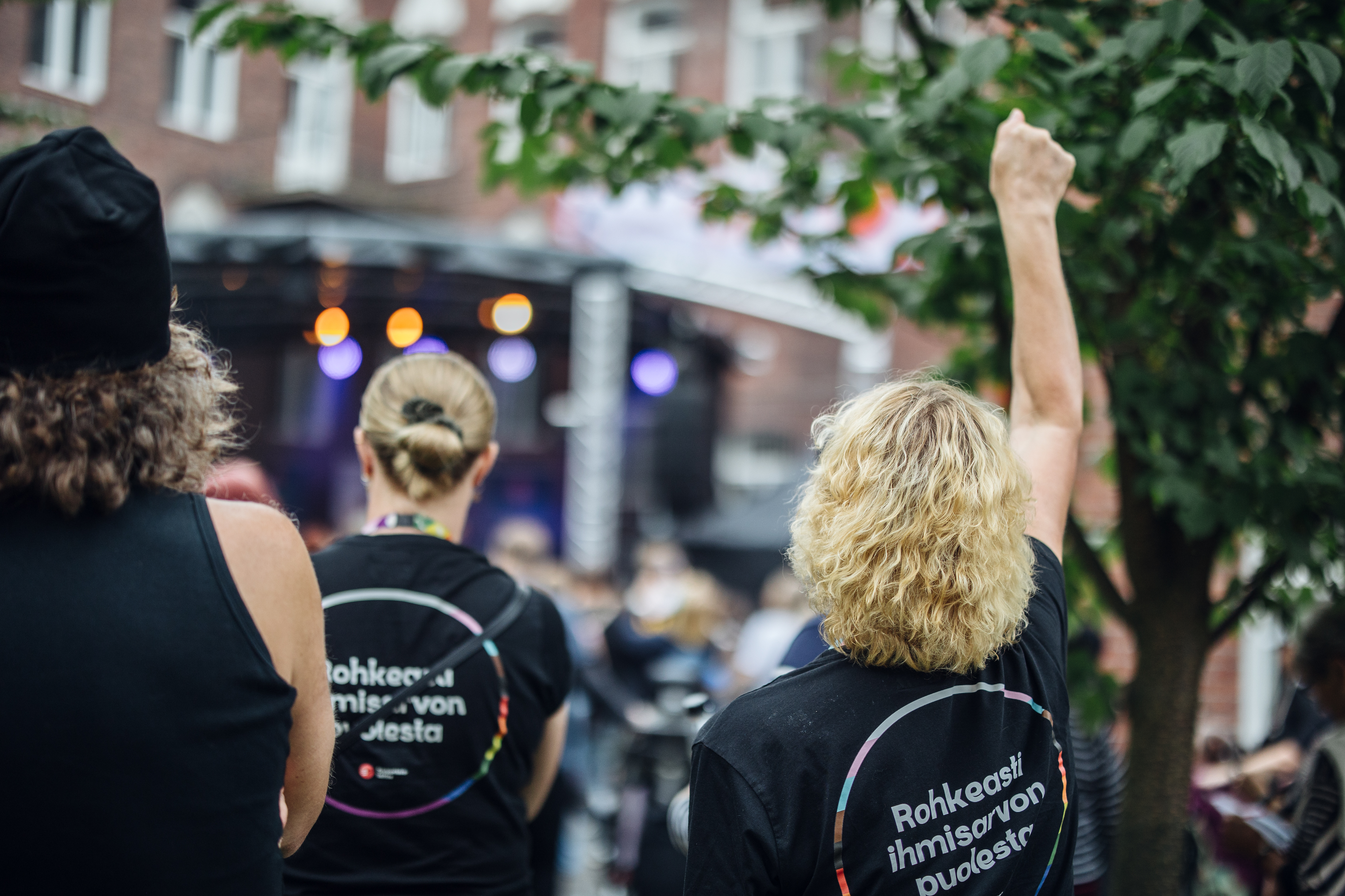 Kolme ihmistä takaa kuvattuna ulkotapahtumassa, yksi nostaa käden ilmaan, heillä on mustat T-paidat, joissa lukee "Rohkeasti ihmisarvon puolesta".