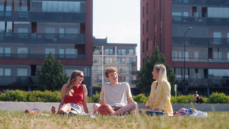 Kolme nuorta istuu nurmikolla keskustellen aurinkoisena päivänä kaupunkimaiseman edustalla.