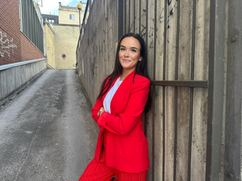 Nuori nainen punaisessa puvussa nojaa puiseen aitaan ulkona kävelykadun vieressä ja hymyilee.