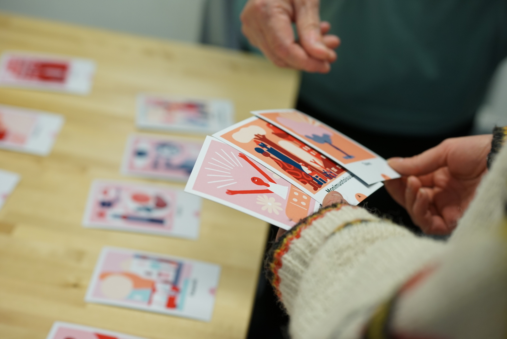 Kädet pitelevät värikkäitä tulevaisuuskortteja pöydän yllä, jossa on lisää kortteja levitettynä, taustalla henkilö seisoo.