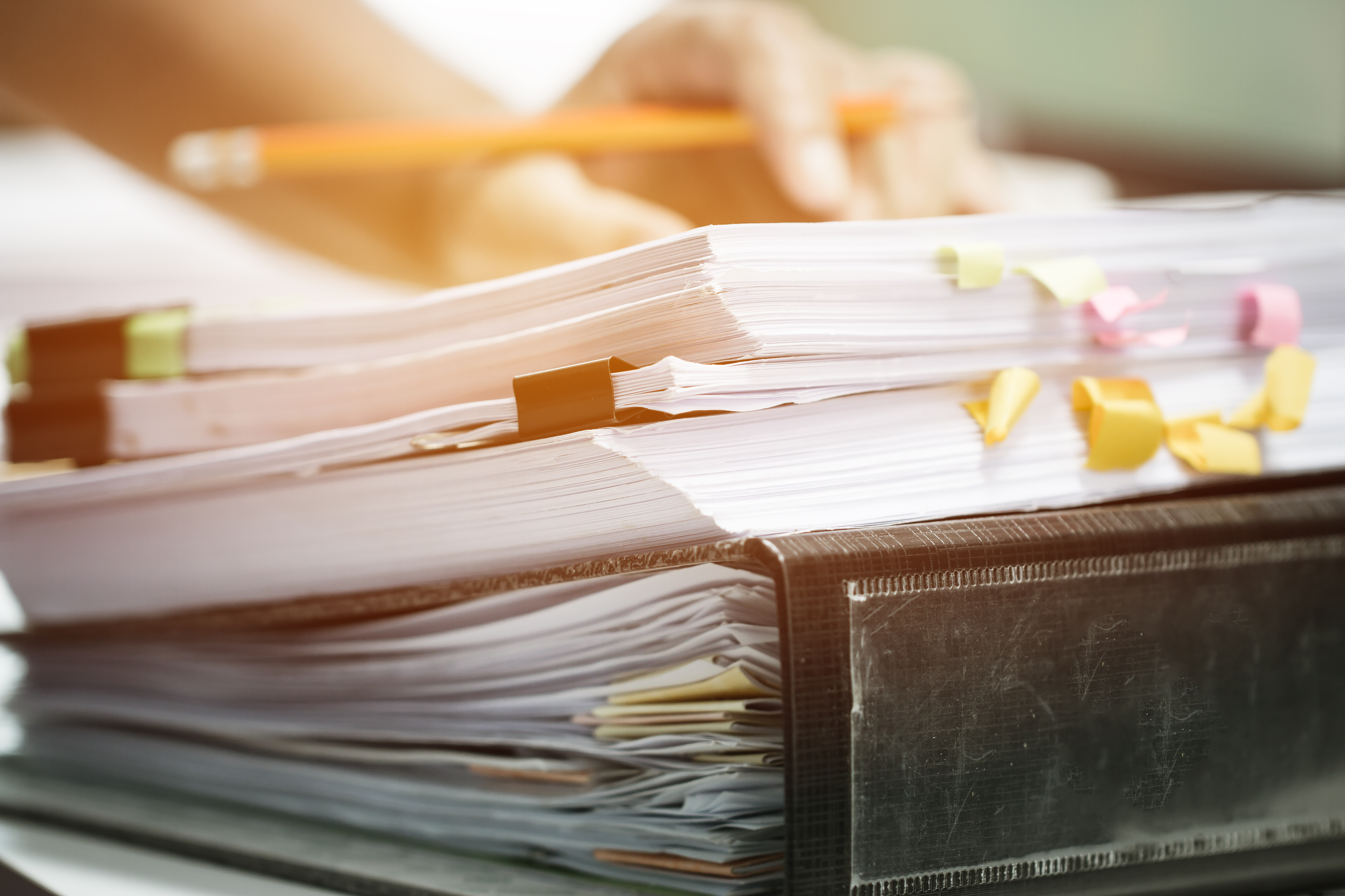 Pino keskeneräisiä asiakirjoja ja raporttitiedostoja toimistopöydällä, taustalla sumeat kädet, jotka kirjoittavat lyijykynällä.