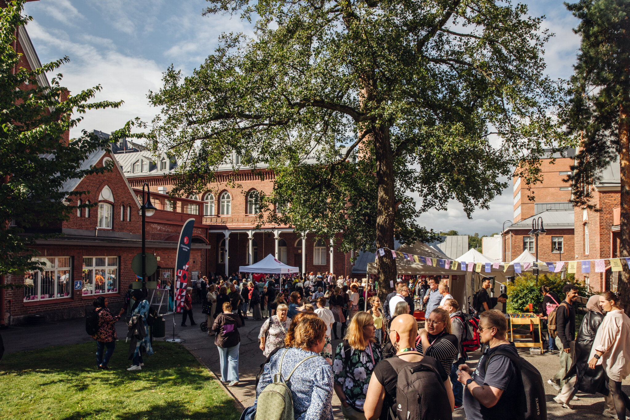 Ihmisiä kokoontuneena ulkoilmatapahtumaan vanhojen punatiilisten rakennusten ympärillä, puita ja sininen taivas.