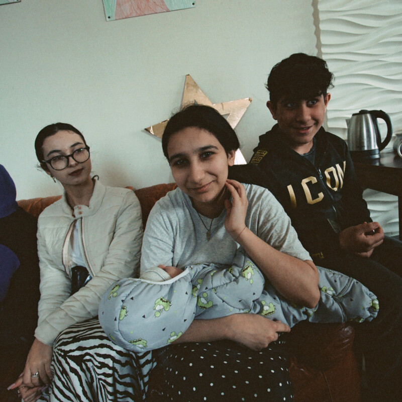 Kolme lasta istuu punaisella sohvalla ja keskimmäinen tyttö pitelee vauvaa sylissään.