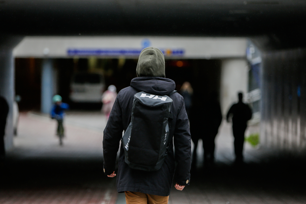 Kuvassa näkyy selin kulkeva henkilö, jolla on reppu ja pipo, kävelemässä hämärässä kaupunkikäytävässä tai tunnelissa, taustalla muita ihmisiä epätarkasti.