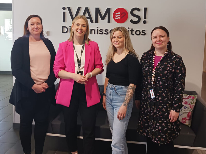 Four women standing side by side smiling inside a building with a sign behind them that reads "¡VAMOS! Diakonissalaitos."