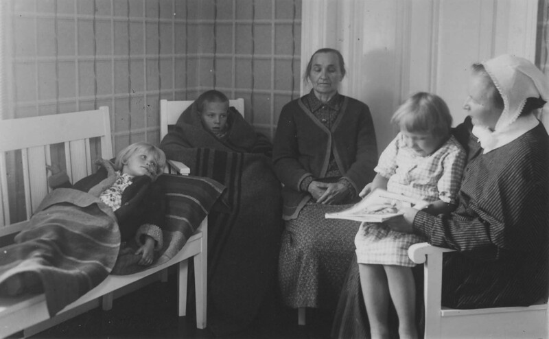 Mustavalkoinen valokuva, jossa diakonissa istuu tuolilla lukemassa lapselle, kolme muuta lasta istuu tai makaa vieressä peittojen alla.