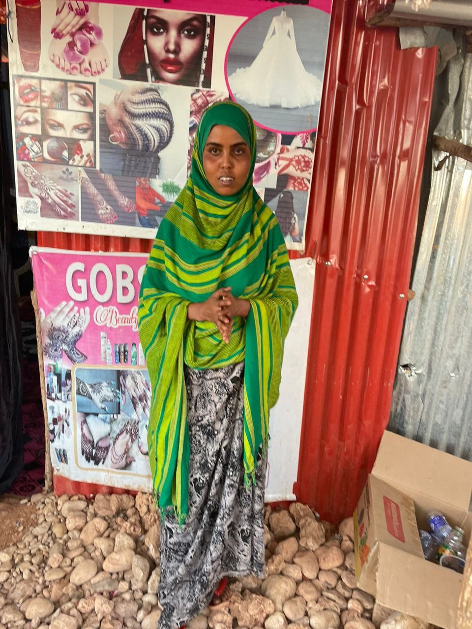 Mekkoon ja suureen vihreään huiviin pukeutunut nuori nainen seisoo aaltopeltiseinään kiinnitettyjen julisteiden edessä.