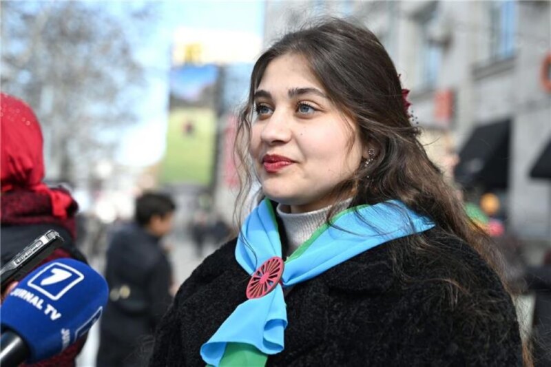 Tummahiuksinen nainen vaaleansininen huivi kaulassaan seisoo kadulla ison mikrofonin edessä.