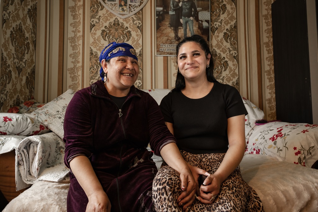 Two women sitting close together on a bed in a warmly decorated room, smiling and holding hands, conveying a sense of support and friendship.