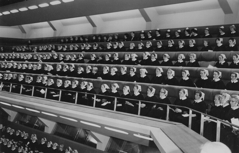 Mustavalkoinen kuva, jossa suuri joukko diakonissoja istuu useilla penkkiriveillä auditorion kaltaisessa tilassa juhlimassa diakonissalaitoksen 100-vuotisjuhlaa.